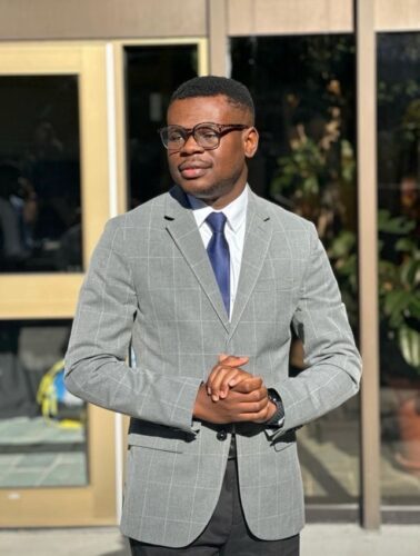 A man in a grey suit jacket and glasses poses for a photo in front of a building.